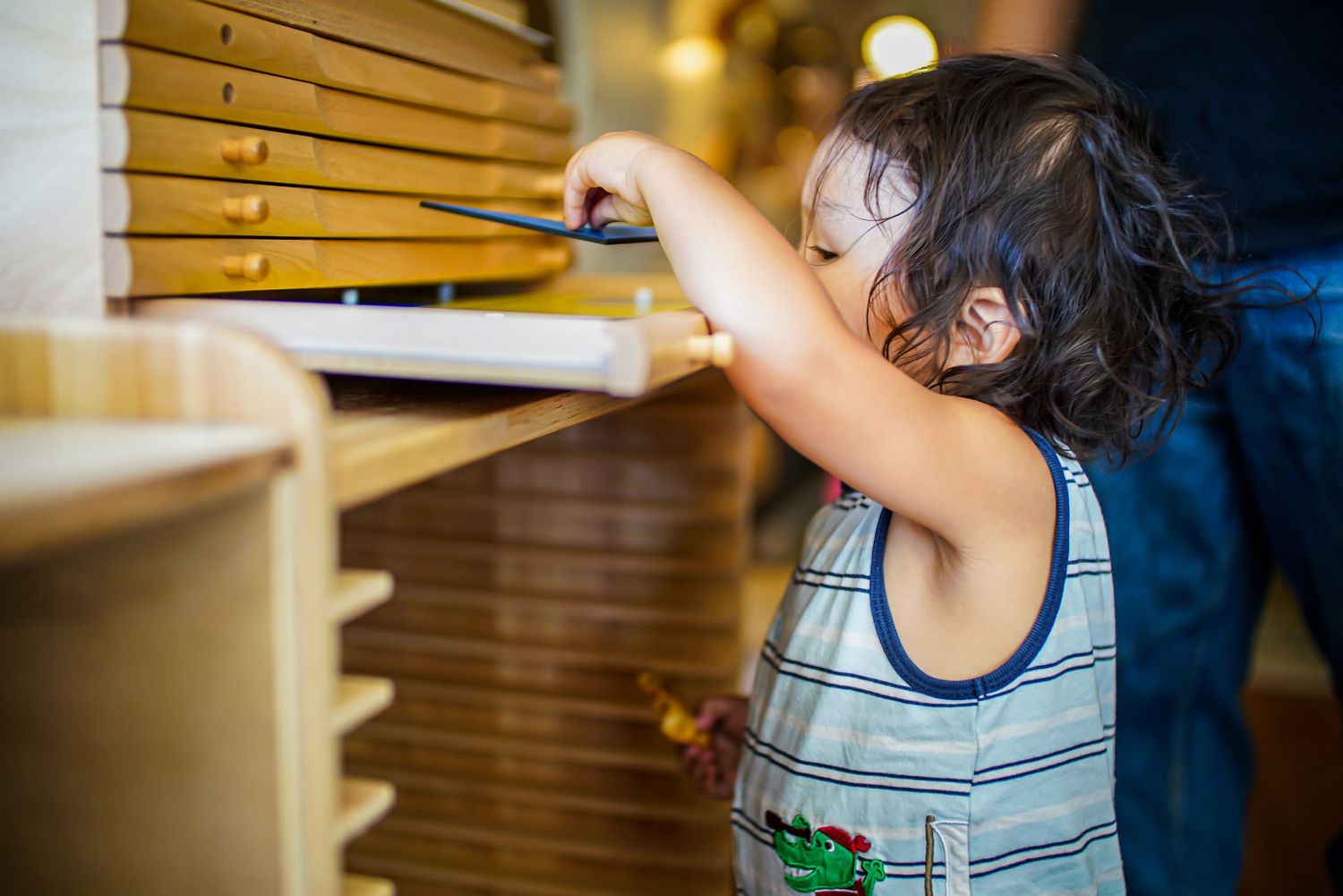 A young child reaching into a wooden flat-file drawer cabinet indoors.