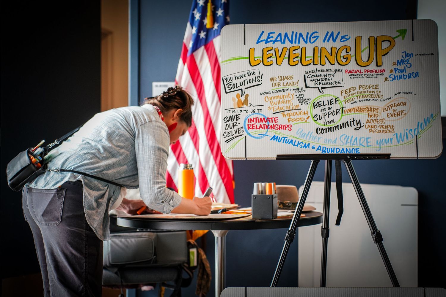 Person leaning over desk writing notes beside colorful Leaning In Leveling Up poster and American flag.