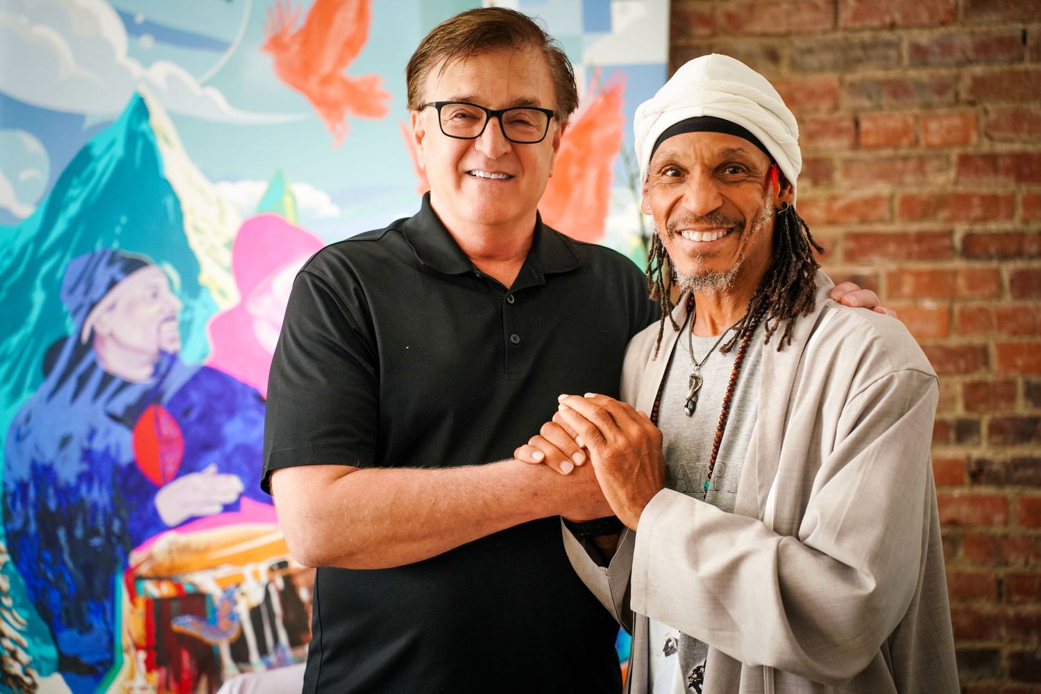 Two smiling men shake hands warmly in front of colorful mural artwork on brick wall.