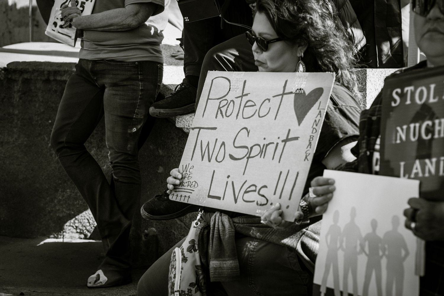 Black and white photo of a woman holding a sign reading Protect Two Spirit Lives at an outdoor protest rally.