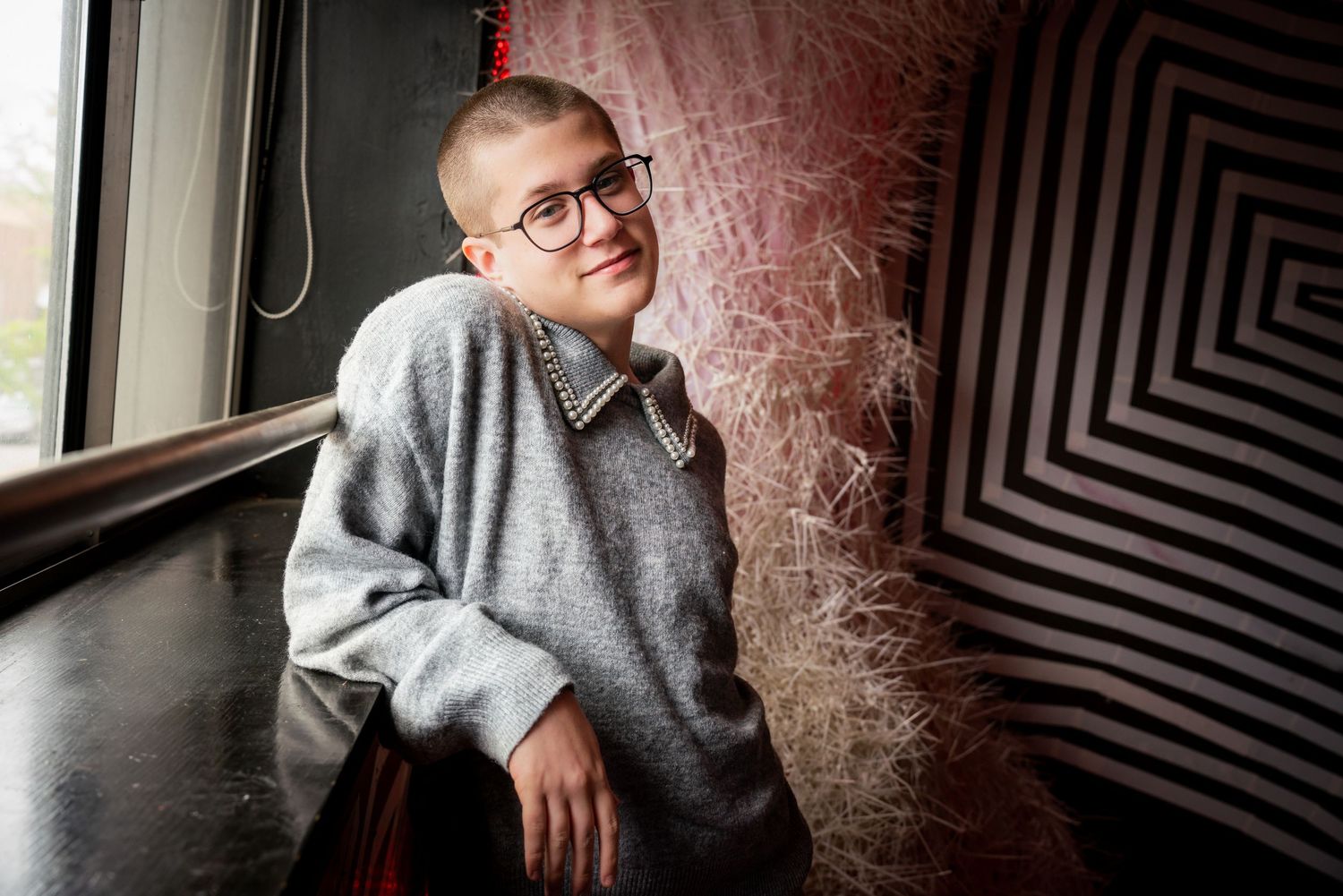 A person with a shaved head wearing a gray sweater and glasses sits near a window by a geometric patterned wall.