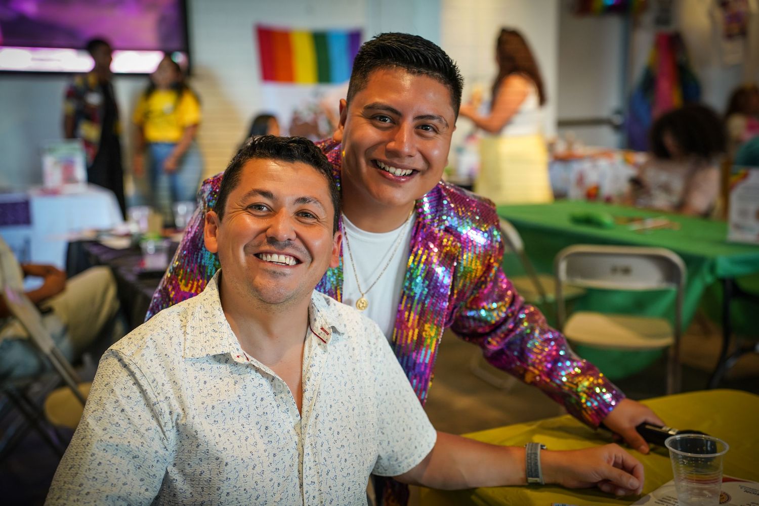 Two smiling men pose together indoors at a colorful Pride celebration event.