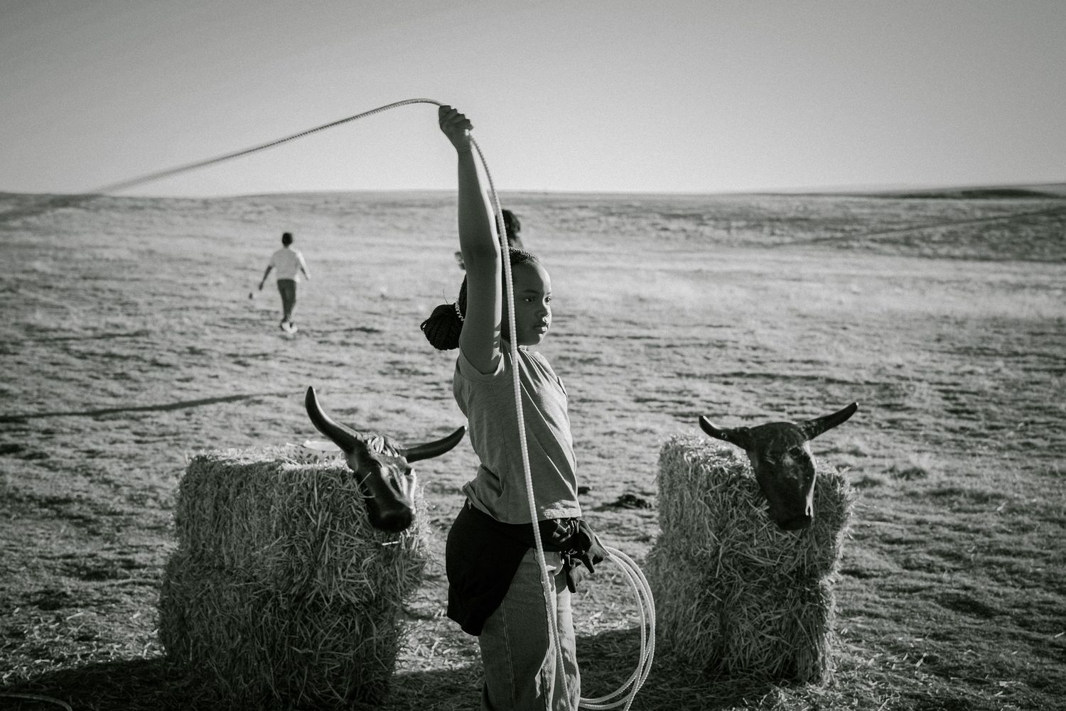 Black and white photo of a person practicing lasso roping over hay bales with bull skulls on a ranch.