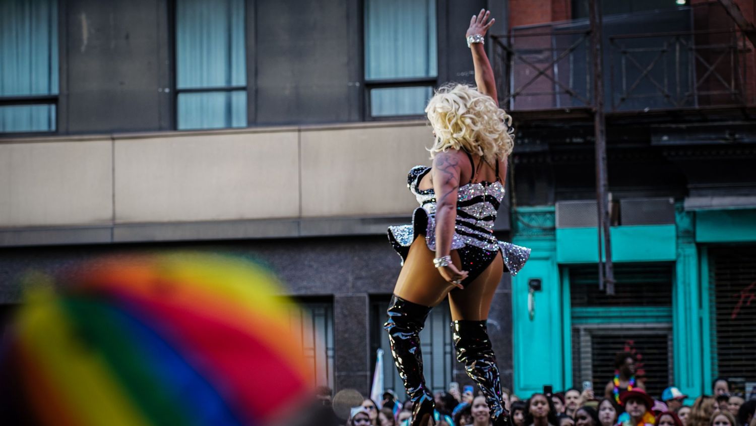 Drag performer in blonde wig and sequined outfit waves to crowd at Pride parade event.