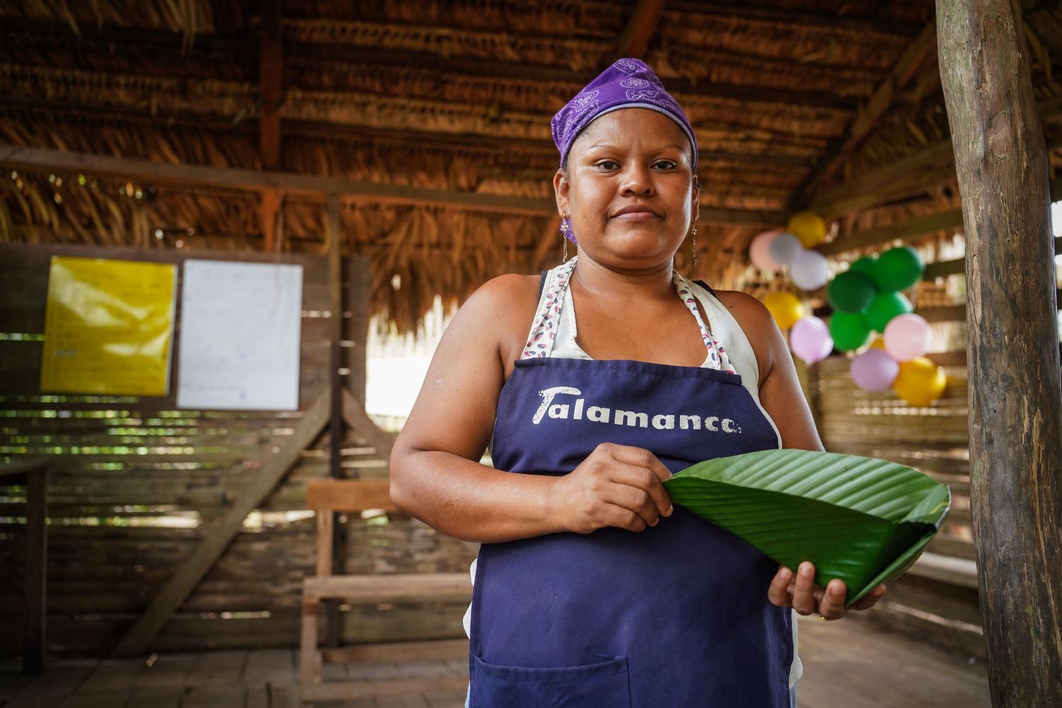 Woman wearing Talamanca apron holding a large green banana leaf in a thatched-roof hut.