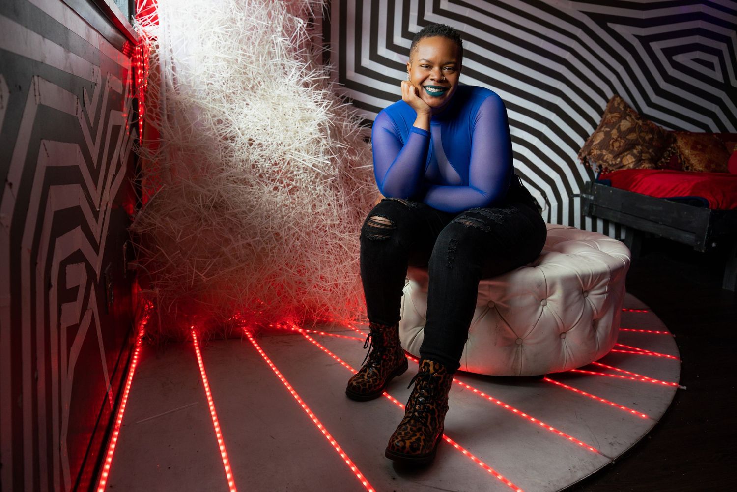 A Black woman in a blue top laughs joyfully while seated on a white ottoman surrounded by red LED lights.