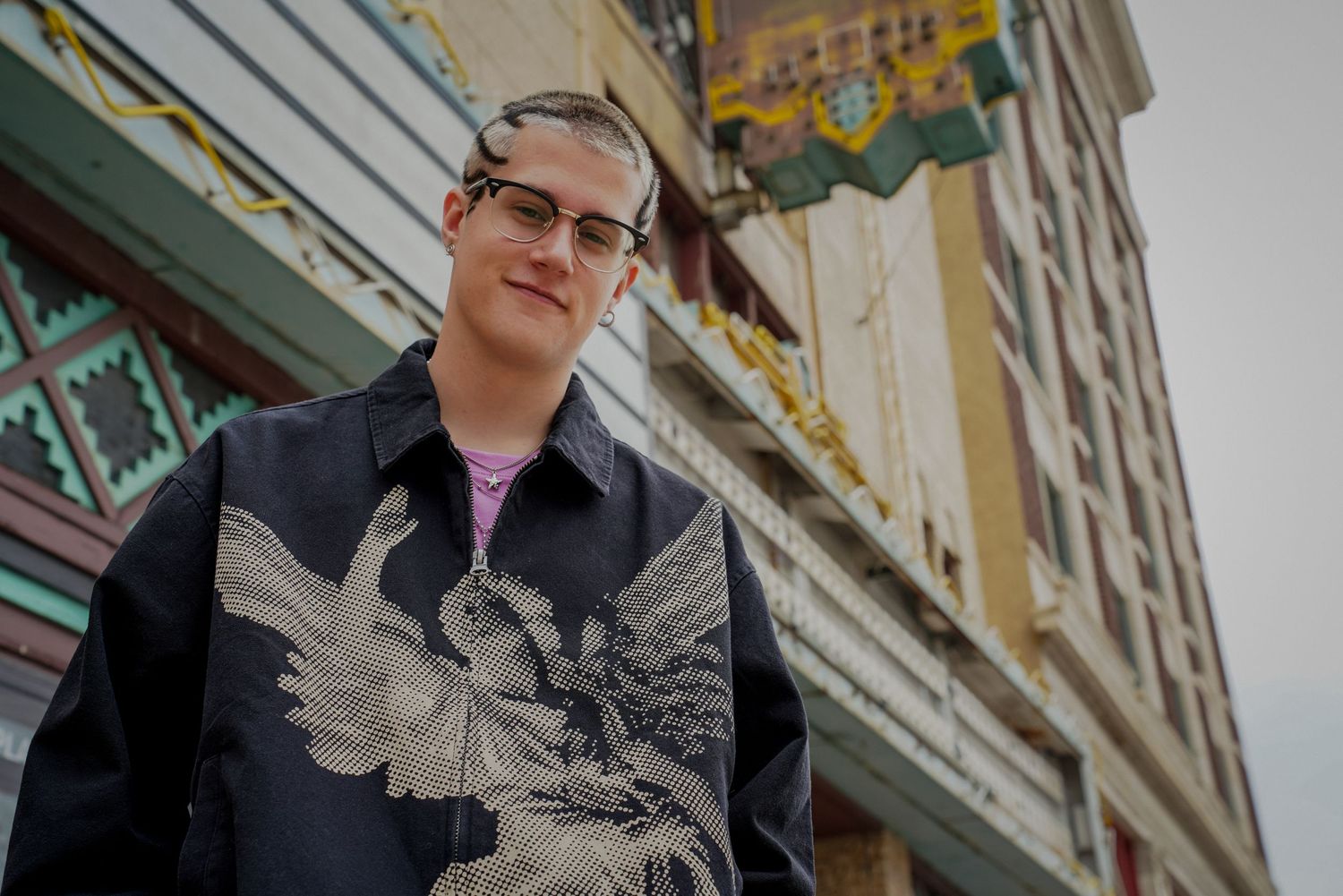 Young man with glasses wearing a black jacket with an embroidered bird design, standing in front of a vintage building with ornate details.