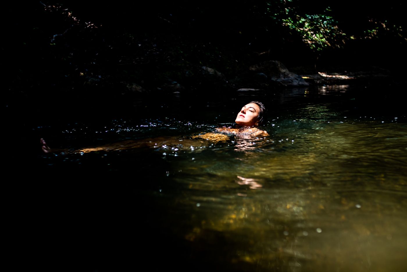 Woman floats peacefully on her back in a sunlit natural forest stream with clear shallow water.