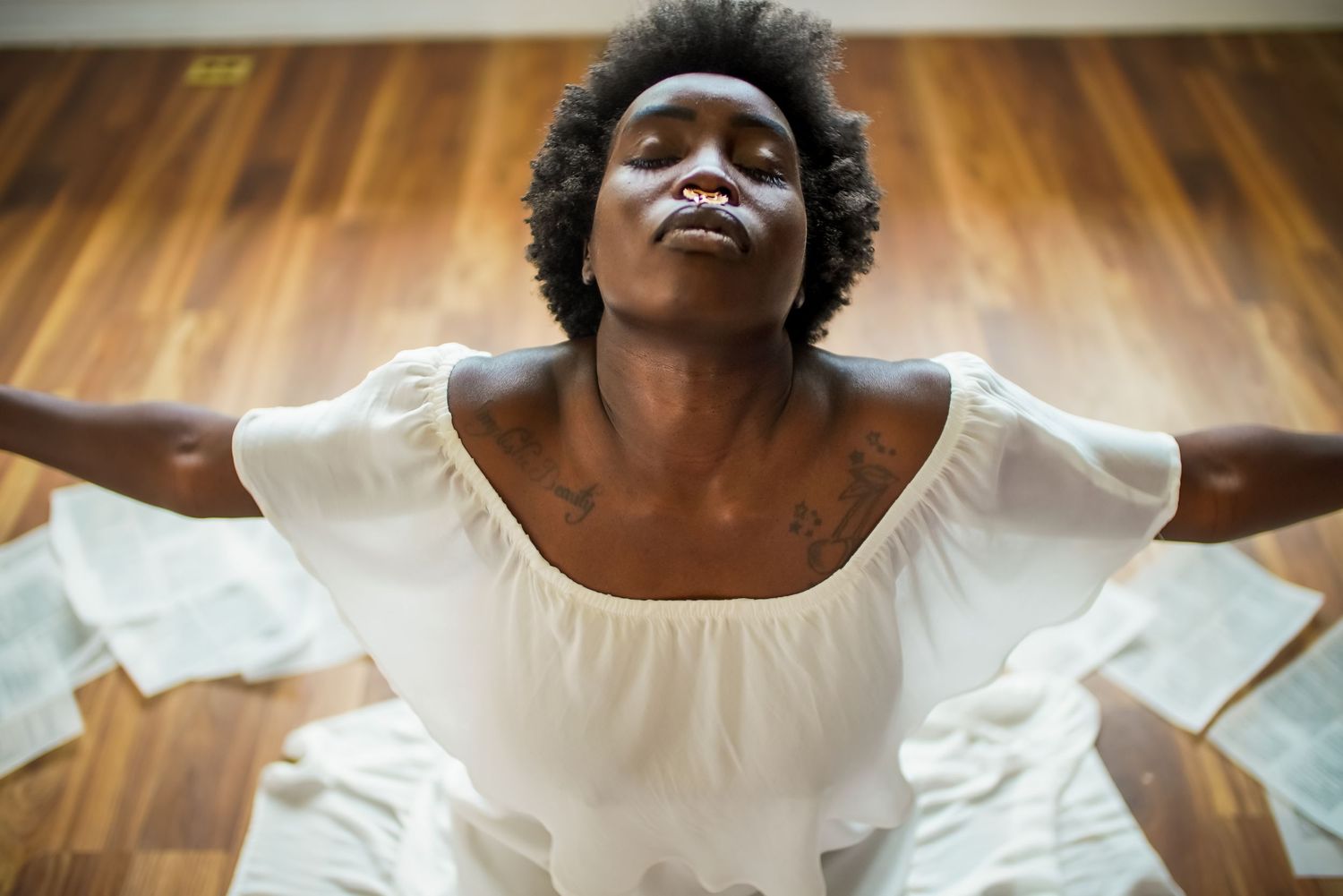 A Black woman in a white dress sits on the floor with arms outstretched and eyes closed in a meditative pose.