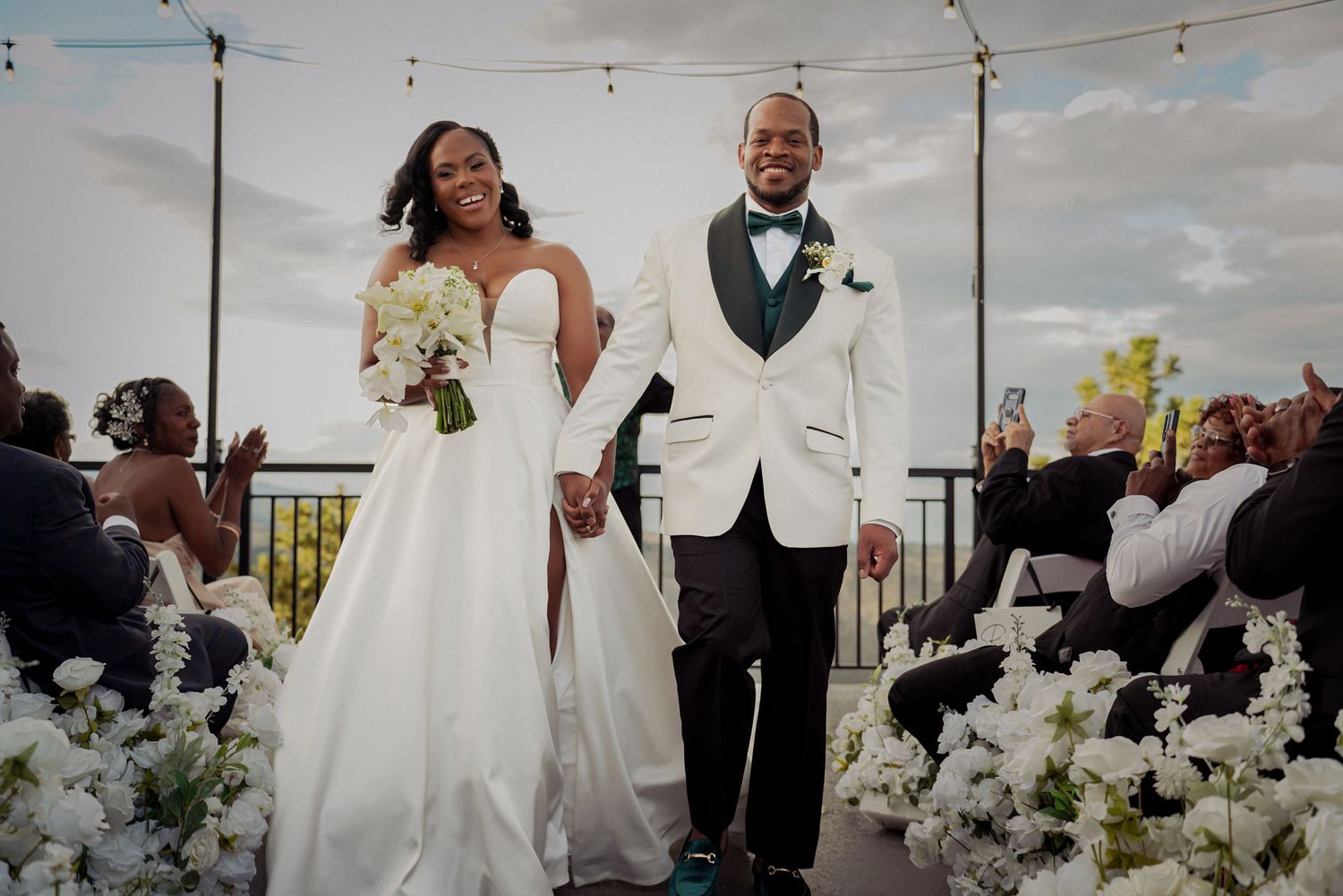 Joyful newlywed couple walking down the aisle at an elegant outdoor wedding ceremony with white floral arrangements.