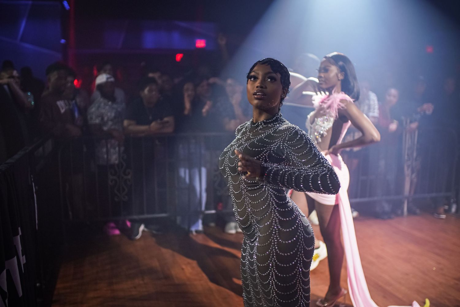 Performer in beaded gown stands on stage under red lights at nightclub fashion event.