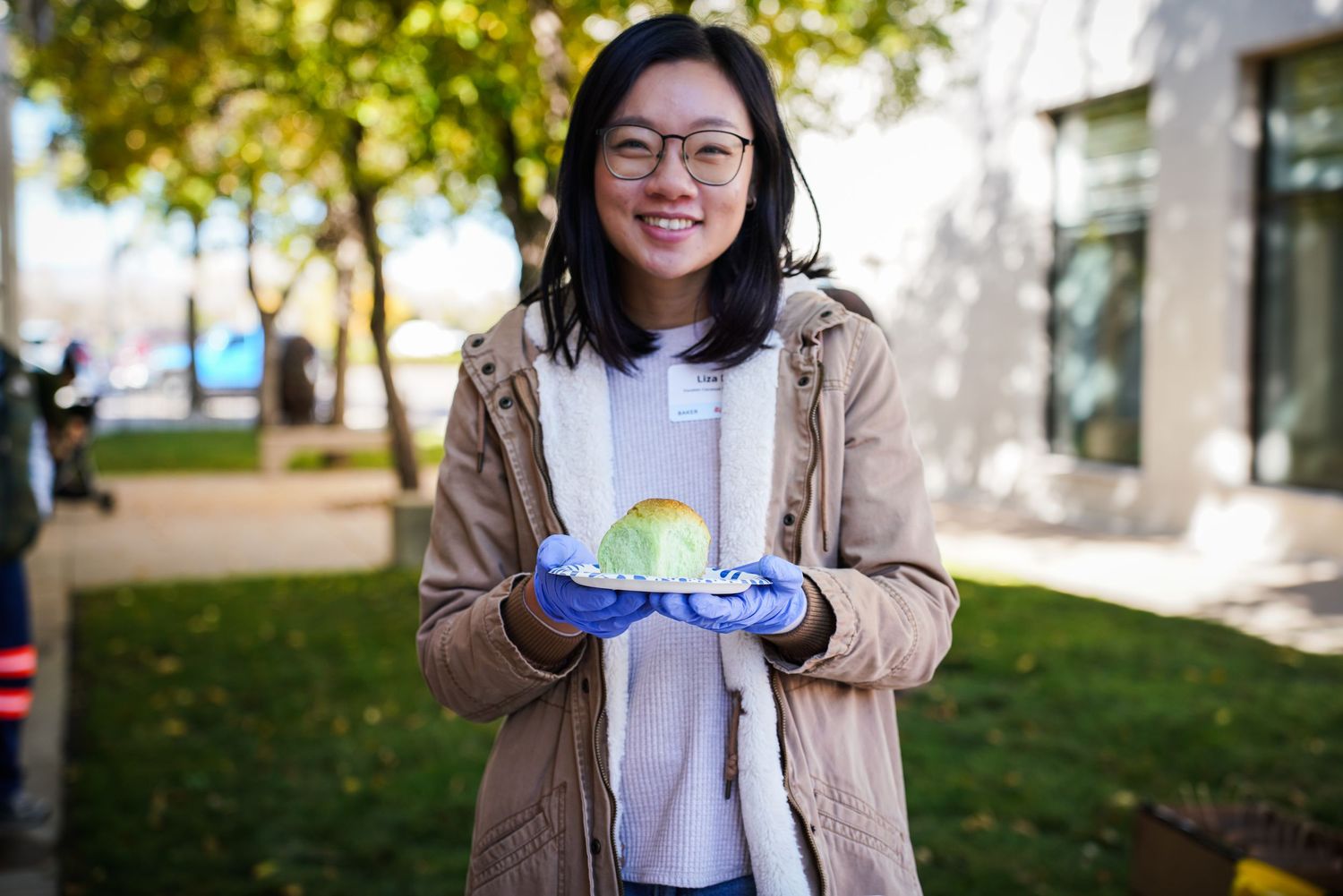 Smiling Asian woman holding a colorful cabbage while wearing blue gloves outdoors.