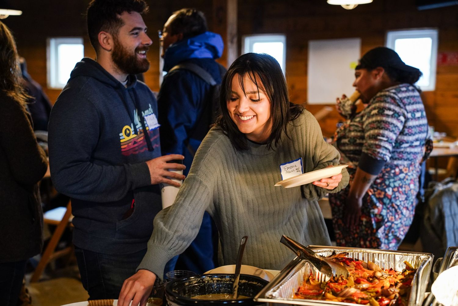 Smiling woman serves herself food at a warm community potluck dinner buffet gathering.