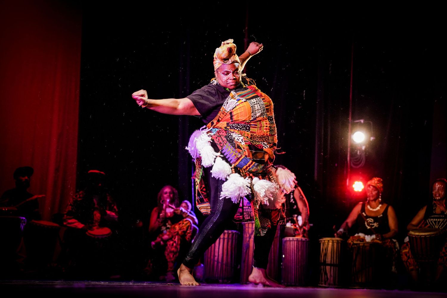 Traditional African dancer performs on stage in colorful costume with feathers under dramatic red lighting.