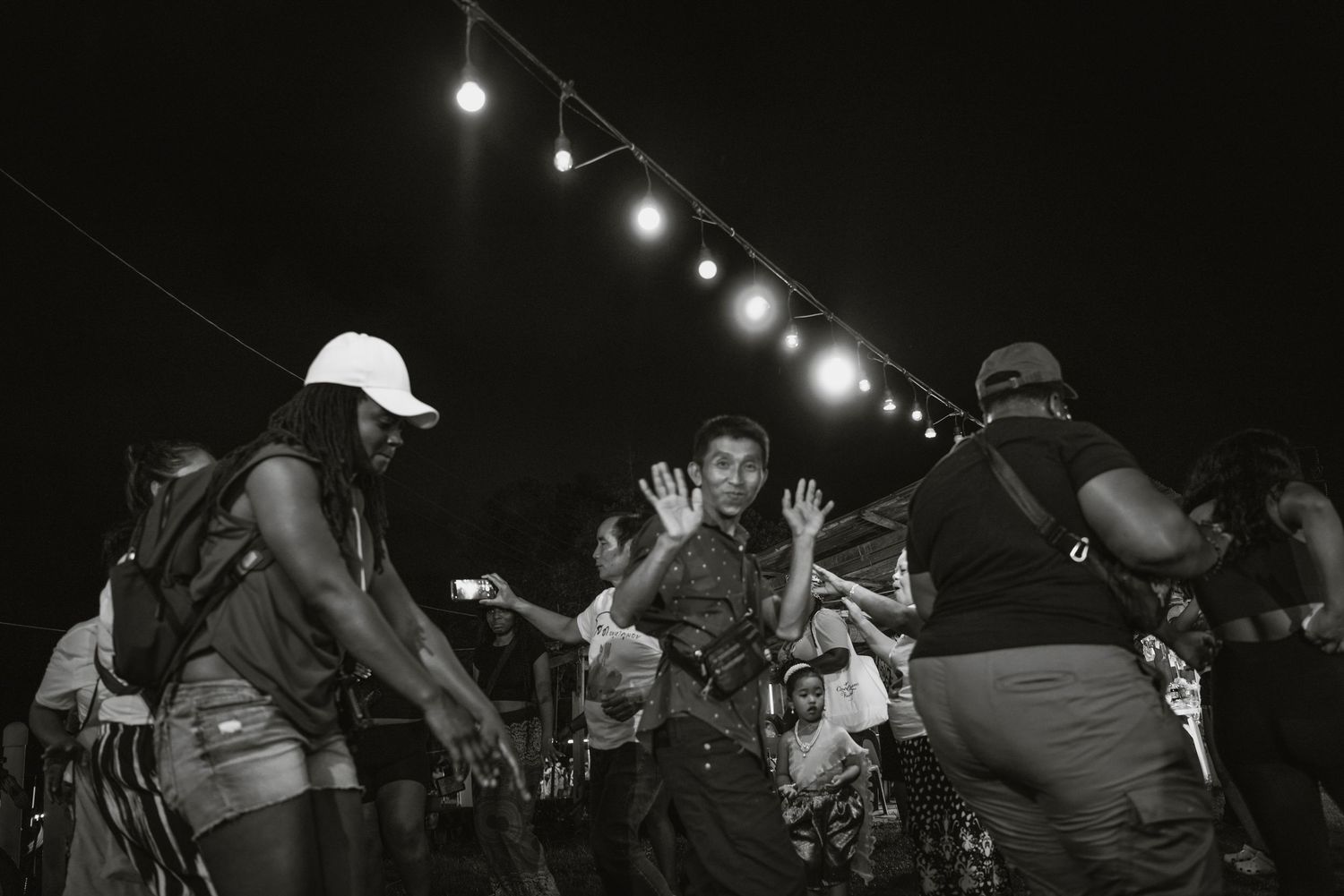 Black and white photo of people dancing outdoors at a night festival under string lights.