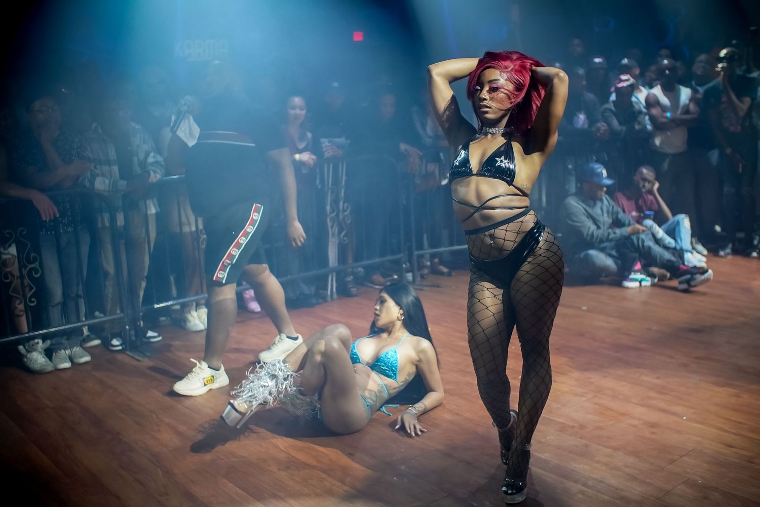 A woman with red hair poses confidently on a dance floor while another performer sits nearby.