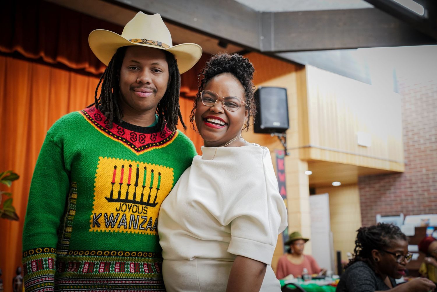 Two women smile together at Kwanzaa celebration, one wearing festive green Kwanzaa sweater and cowboy hat.