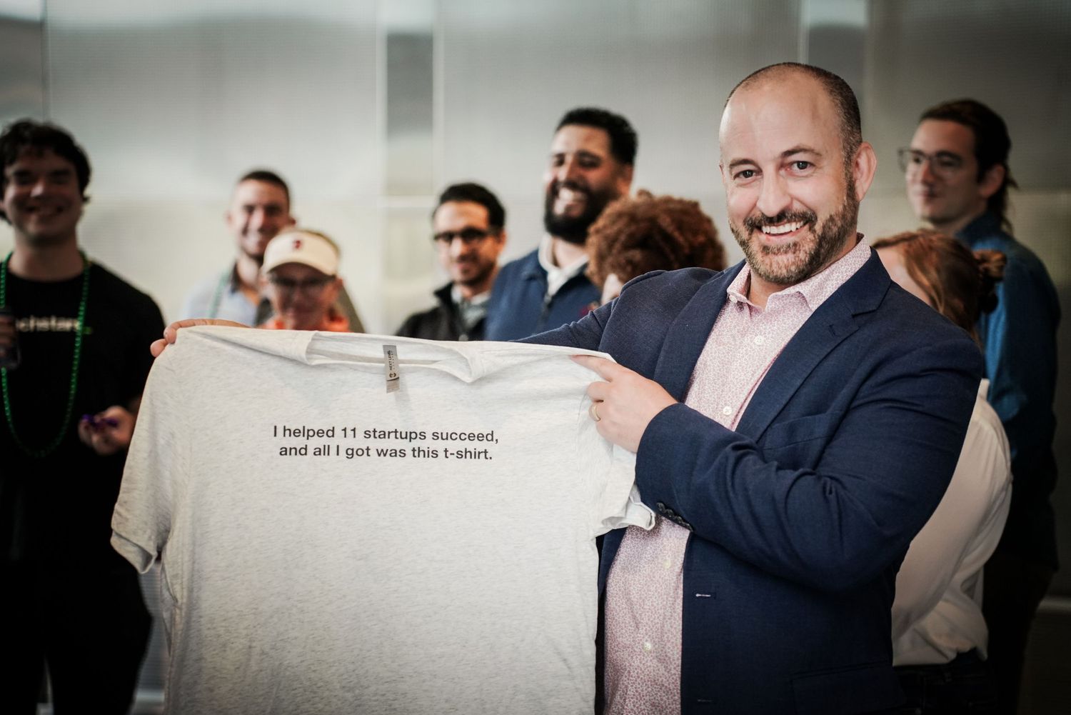 Smiling man holds gray t-shirt reading startup success quote at entrepreneurship networking event.