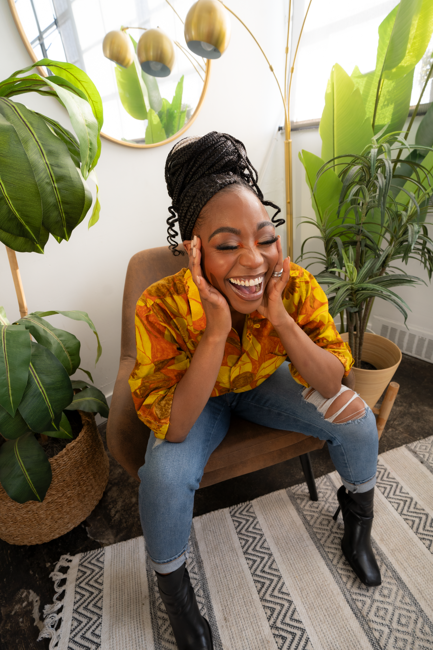 Joyful Black woman in yellow African print shirt sitting in chair surrounded by houseplants.