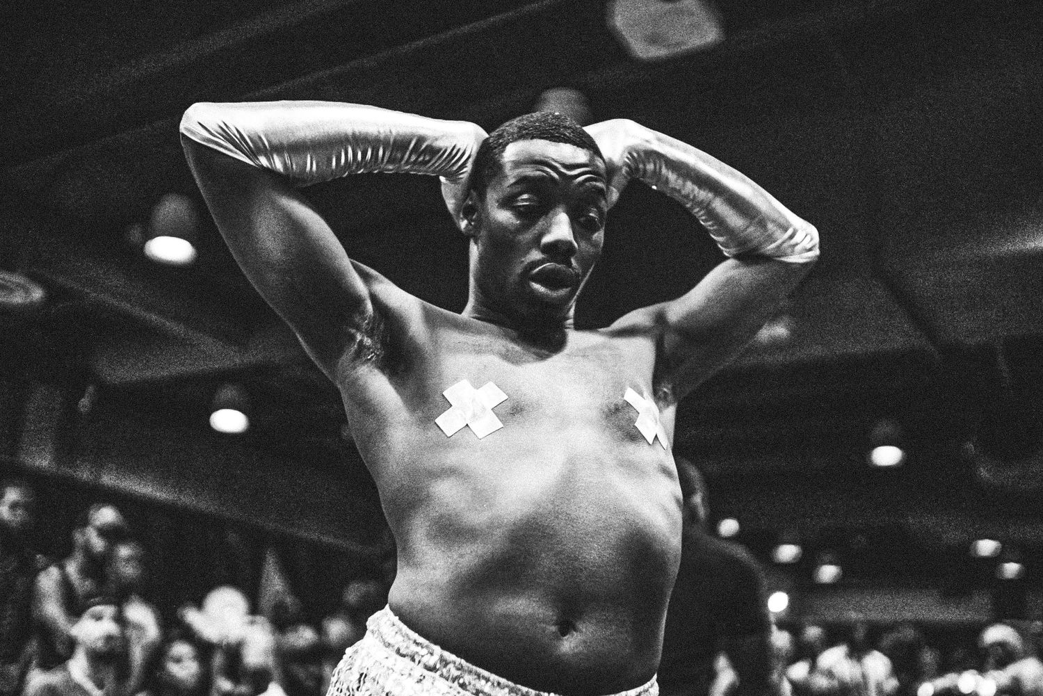 Black and white portrait of shirtless male performer with cross tape on chest at energetic live event.