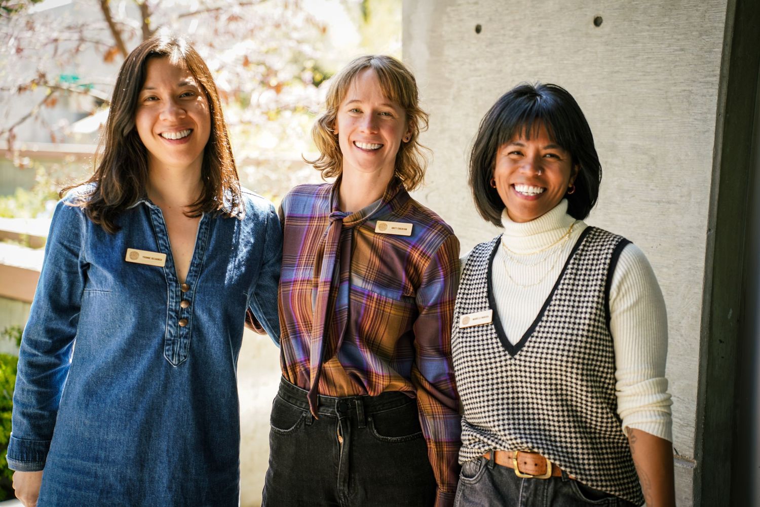 Three smiling women with name tags pose together outdoors near blooming cherry blossom tree.