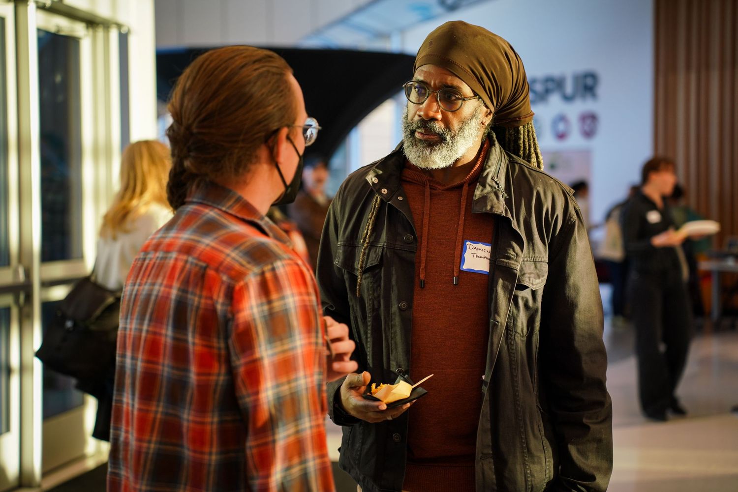 Two men engaged in conversation at networking event, one wearing traditional Sikh turban and holding food.