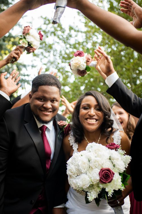 Smiling newlyweds walking under raised bouquets at outdoor wedding celebration.