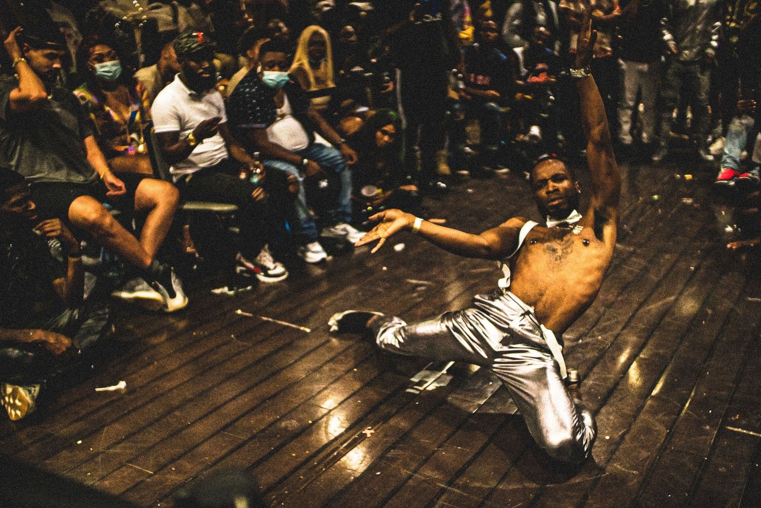 A shirtless dancer performs a floor move in silver pants at an energetic dance competition.