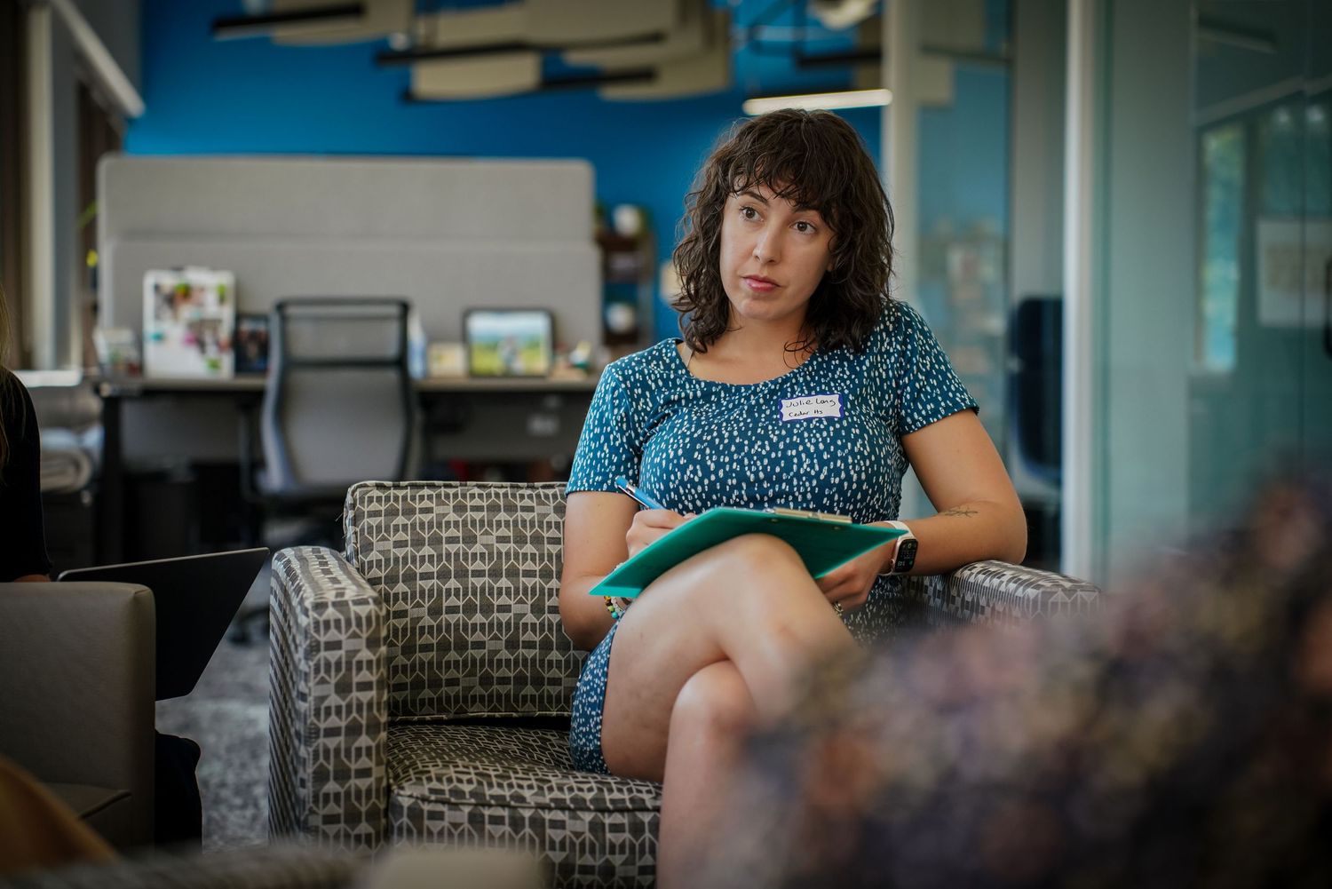 Young woman sitting in a patterned chair holding a notebook in a modern office space.