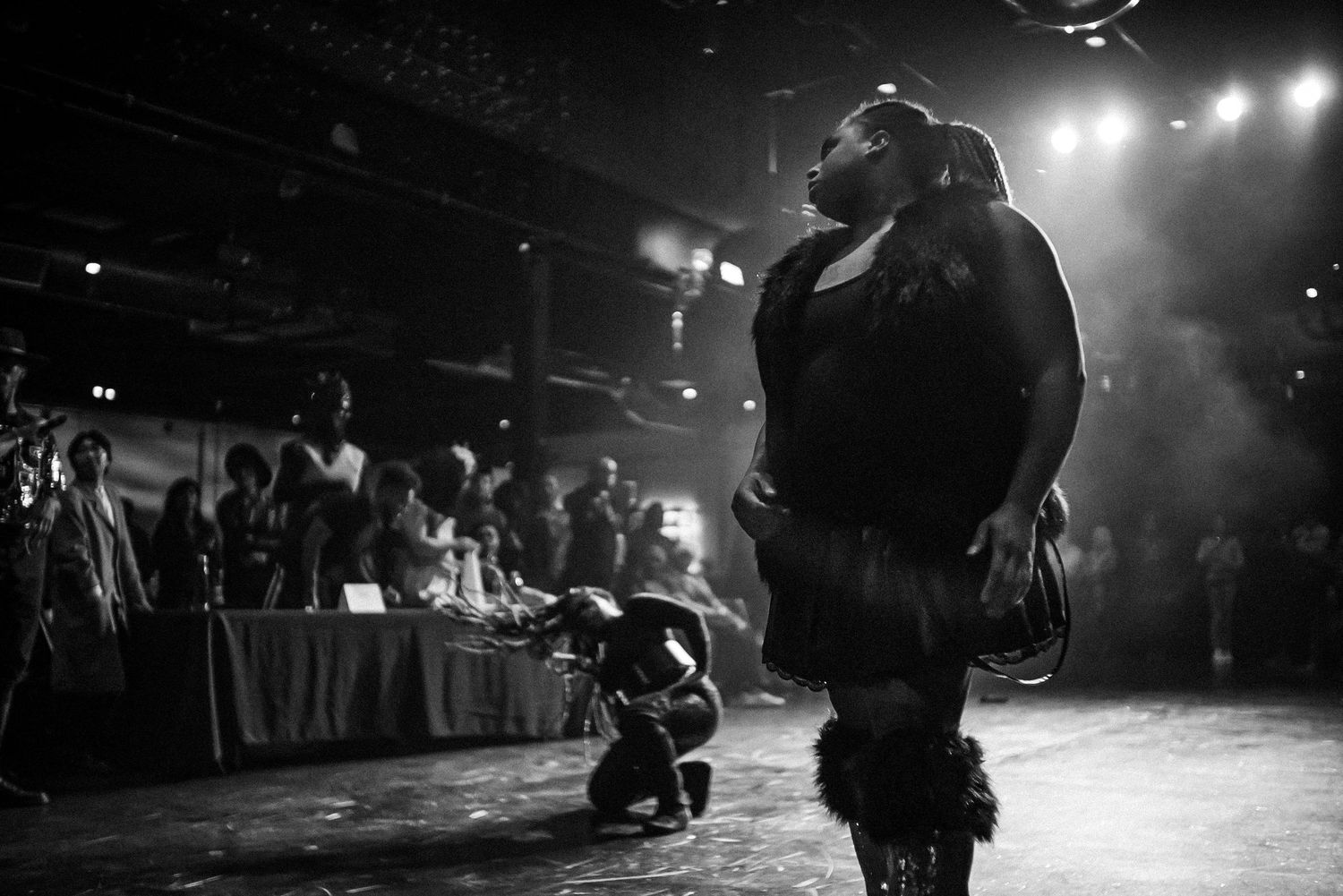 Black and white photo of a plus-size performer on stage under spotlight at a packed nightclub.