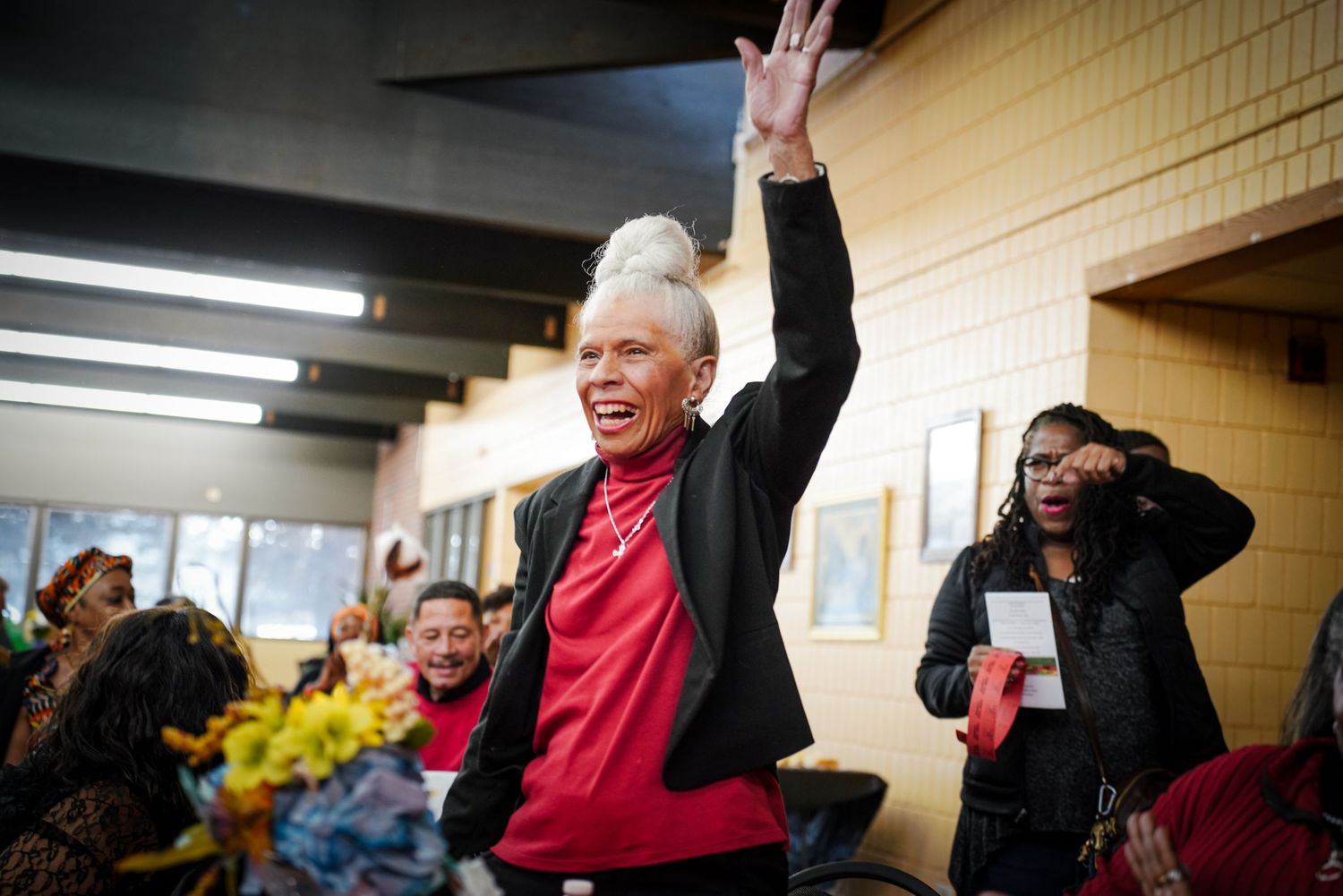 Joyful woman in red outfit raises hand during a lively Kwanzaa cultural celebration indoors.