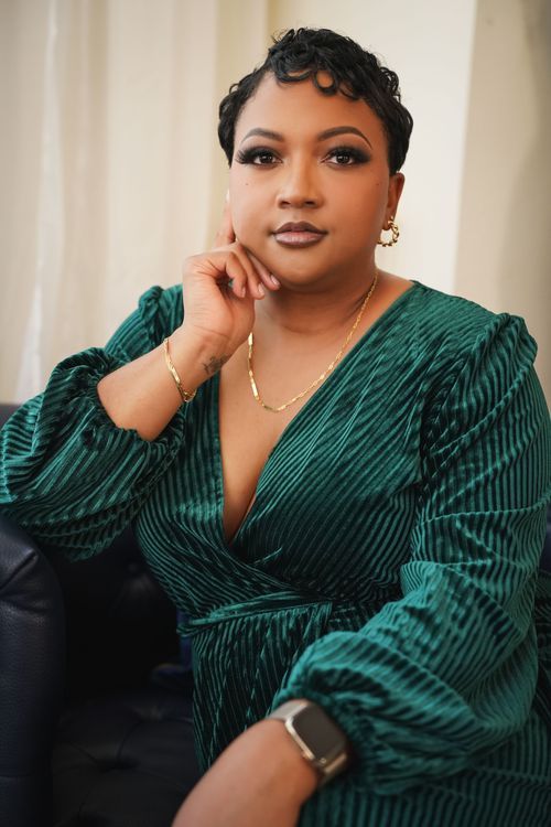 Woman in green velvet wrap dress poses confidently seated, wearing gold jewelry and smartwatch.