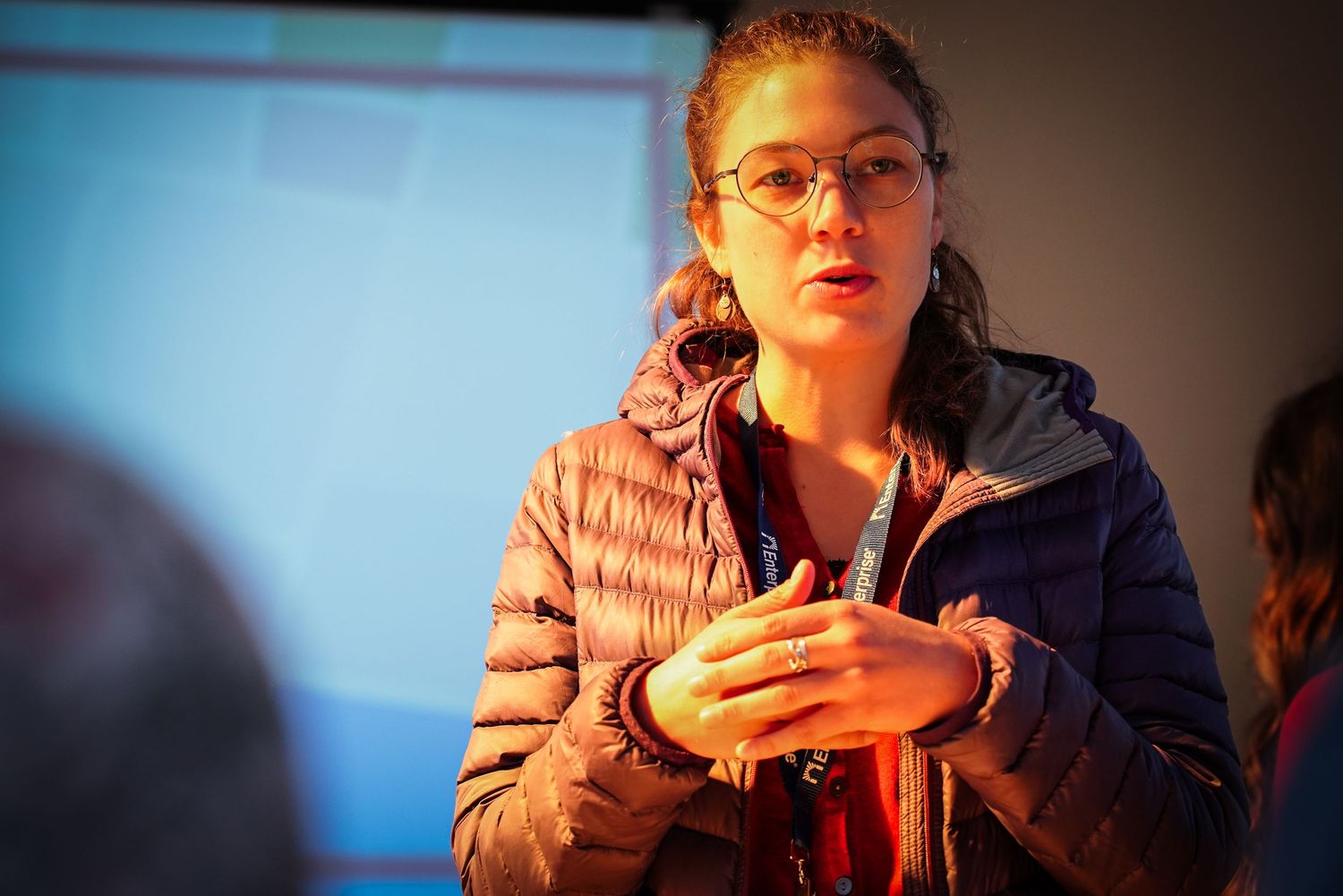 Young woman with glasses speaks expressively while wearing puffer jacket in warmly lit indoor setting.