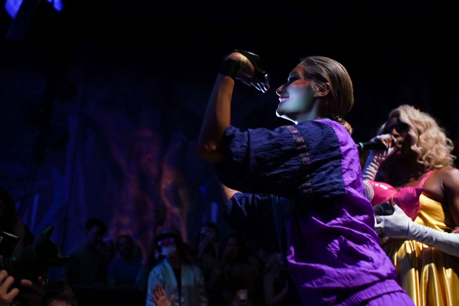 Two performers sing into microphones on colorful lit stage, one in purple jacket, one in gold dress.