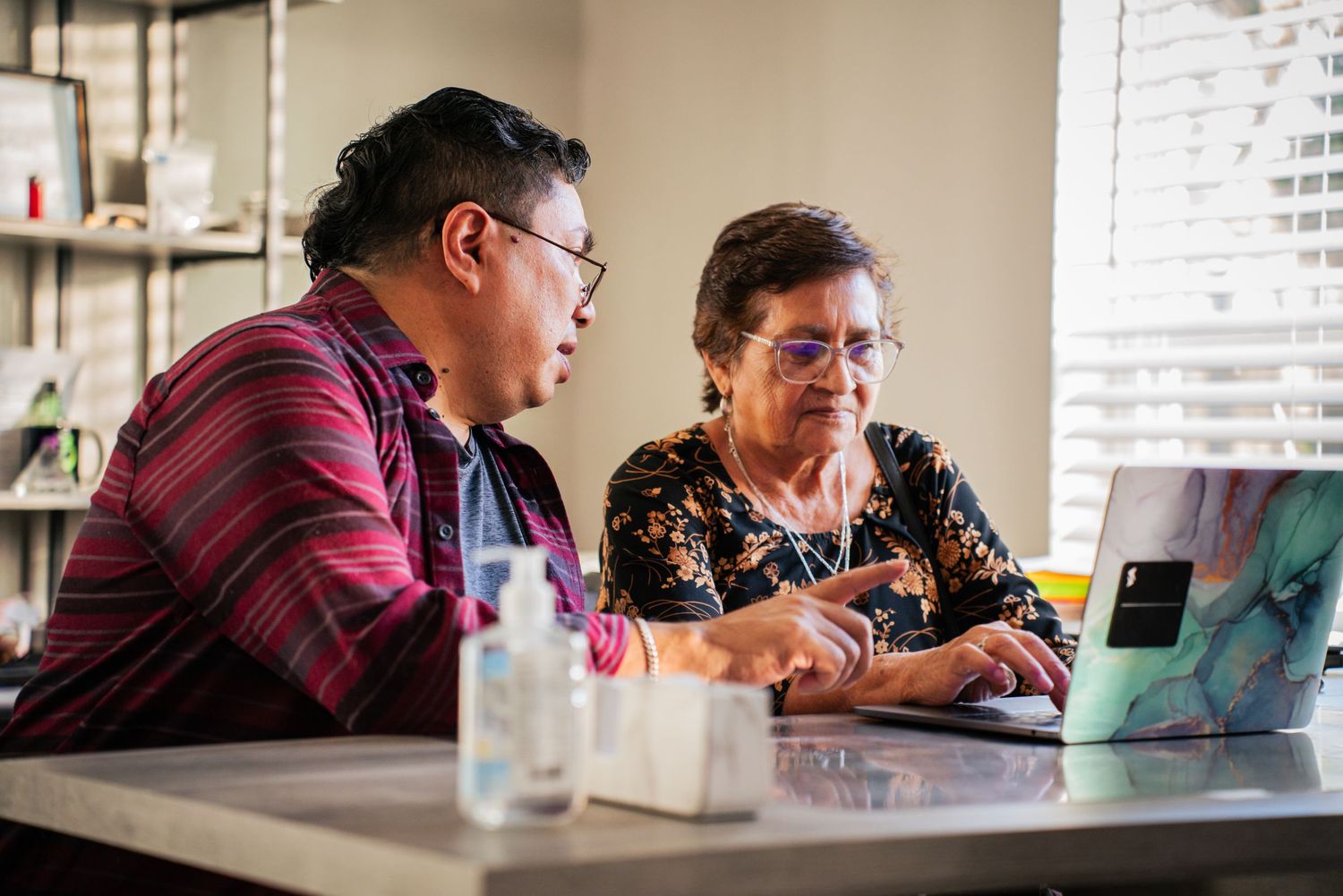 Young man helps elderly woman use a laptop computer at a bright kitchen counter.