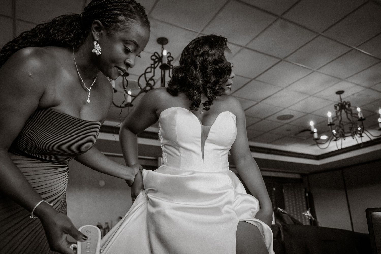 Black and white photo of a bridesmaid helping a bride in a strapless wedding gown prepare in an elegant venue.