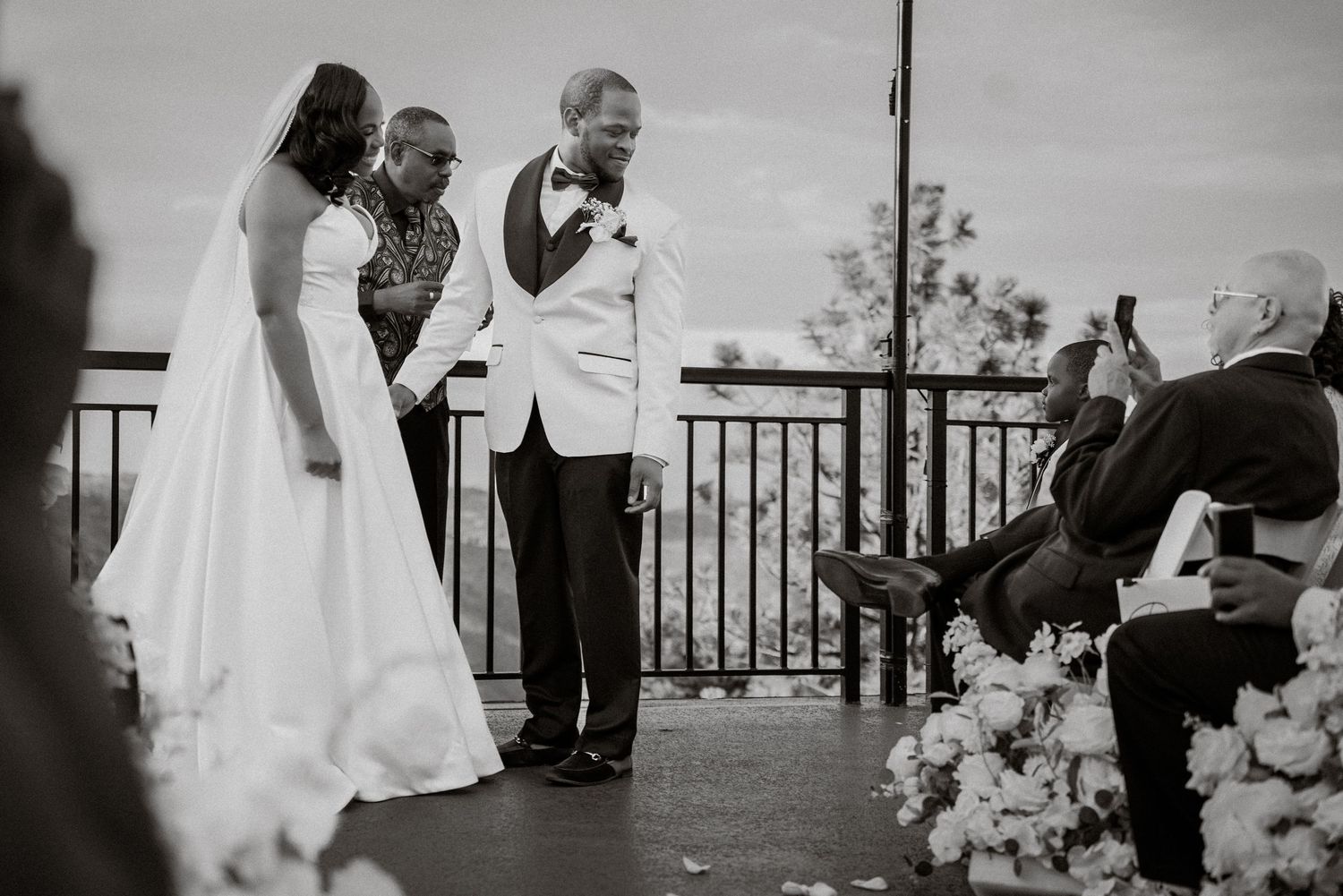 Black and white wedding ceremony photo of bride and groom holding hands at outdoor altar with floral arrangements.