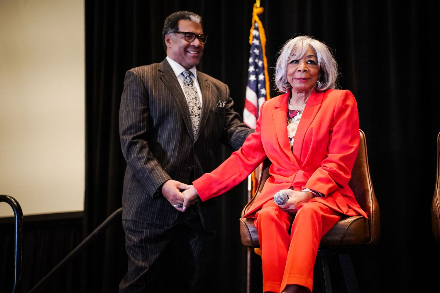 Man in suit stands beside woman in red pantsuit holding microphone on stage with American flag backdrop.