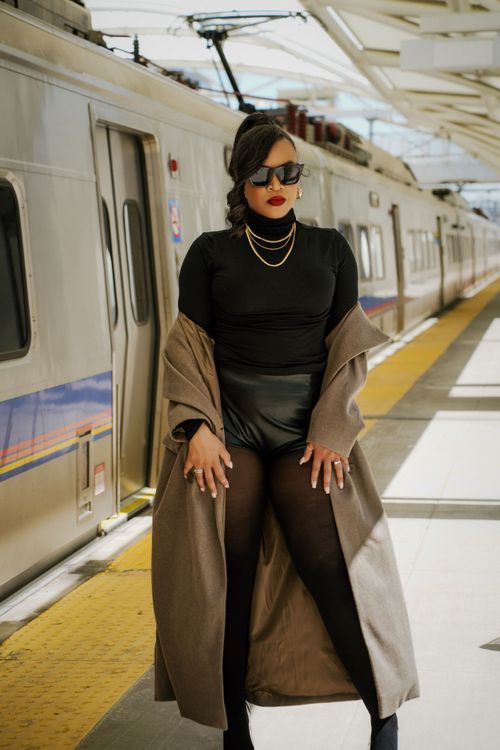 Stylish woman in black outfit and trench coat posing at train platform.