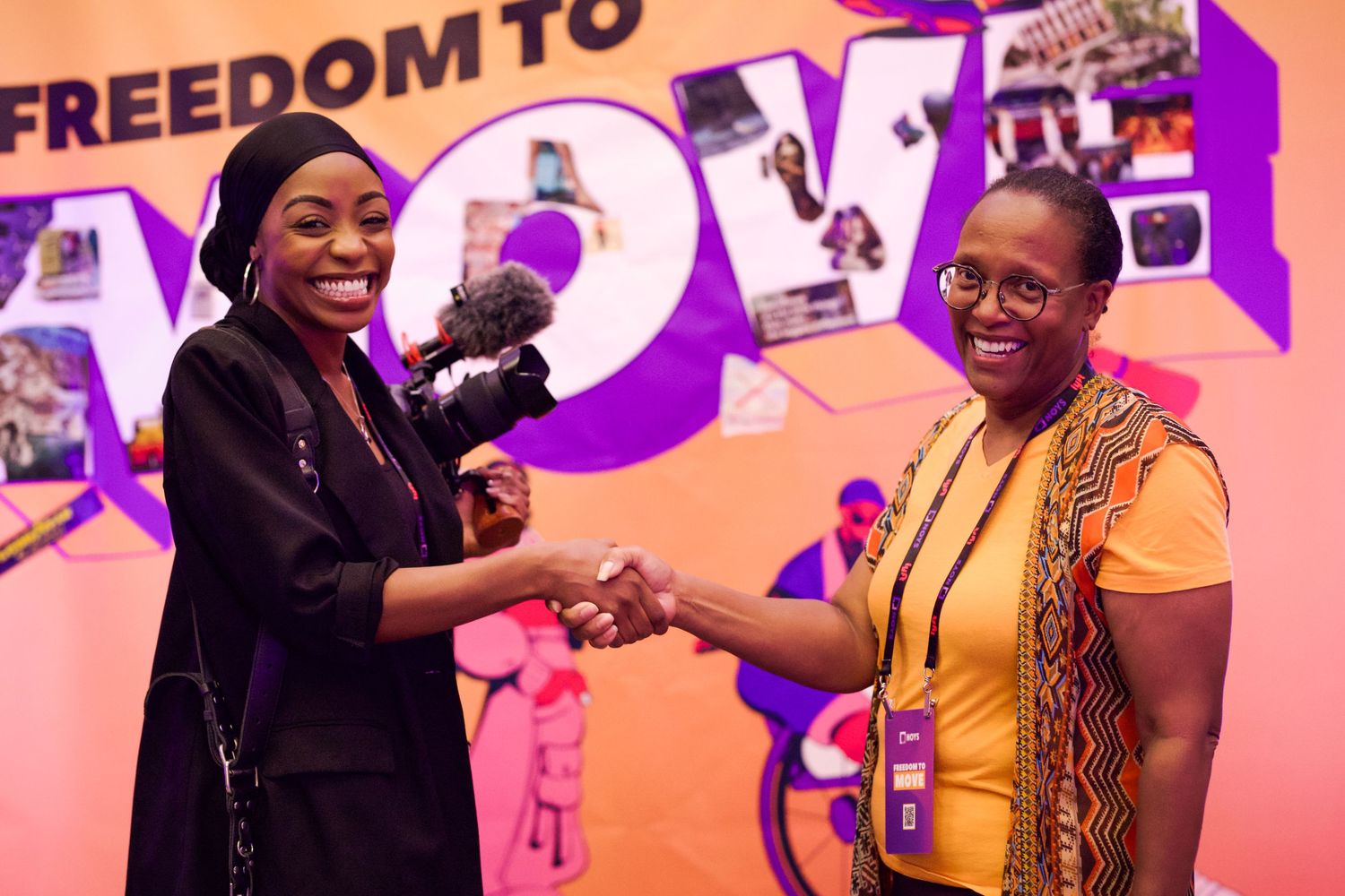 Two women shaking hands in front of a colorful Freedom to Move event backdrop with a video camera.