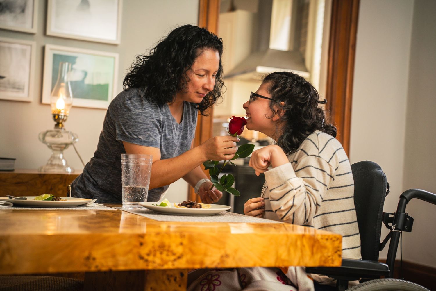 Mother offers a red rose to her daughter in a wheelchair during a warm family meal at home.