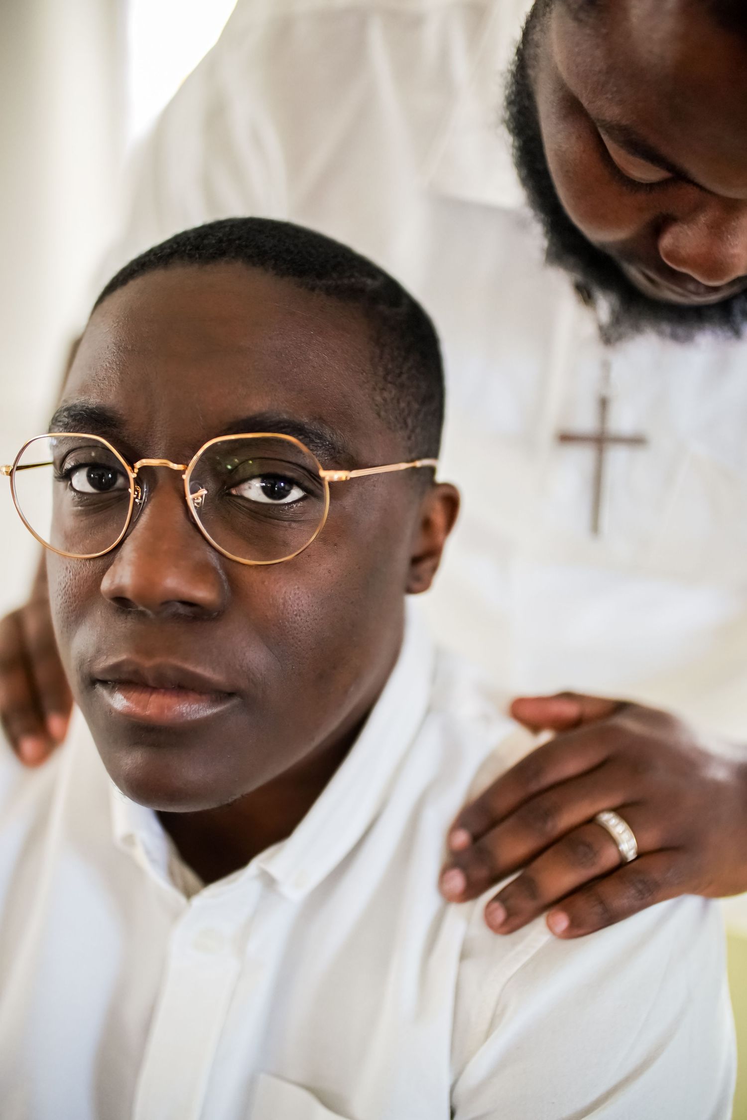 A young Black man wearing glasses and white shirt sits while a religious figure in white robes places hands on his shoulders.