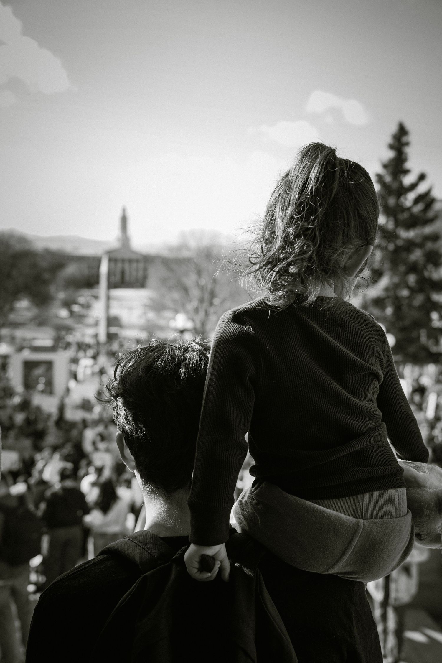 Black and white photo of a child sitting on an adults shoulders overlooking a large outdoor protest crowd.