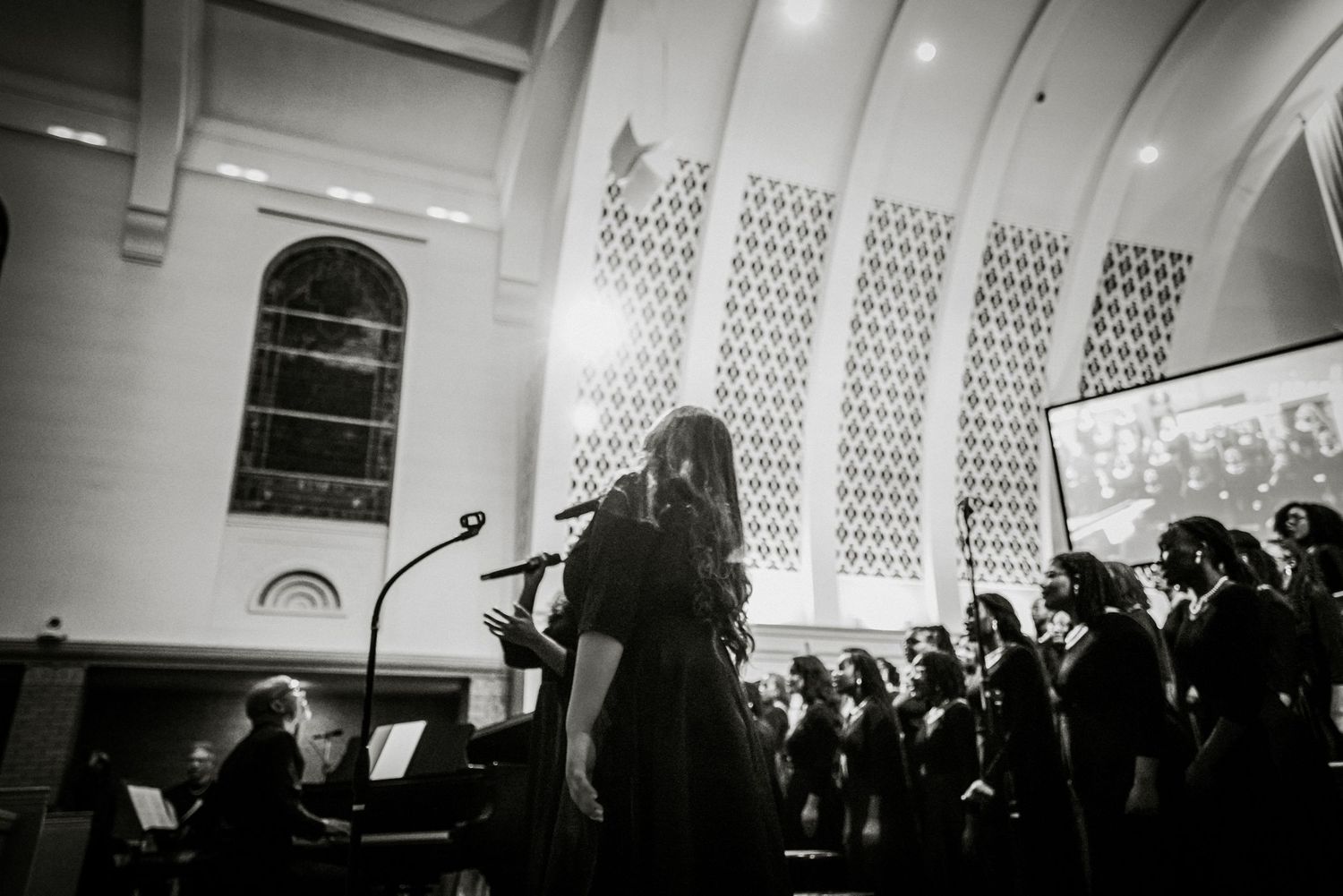 Black and white photo of female soloist performing with choir in elegant church with arched windows.