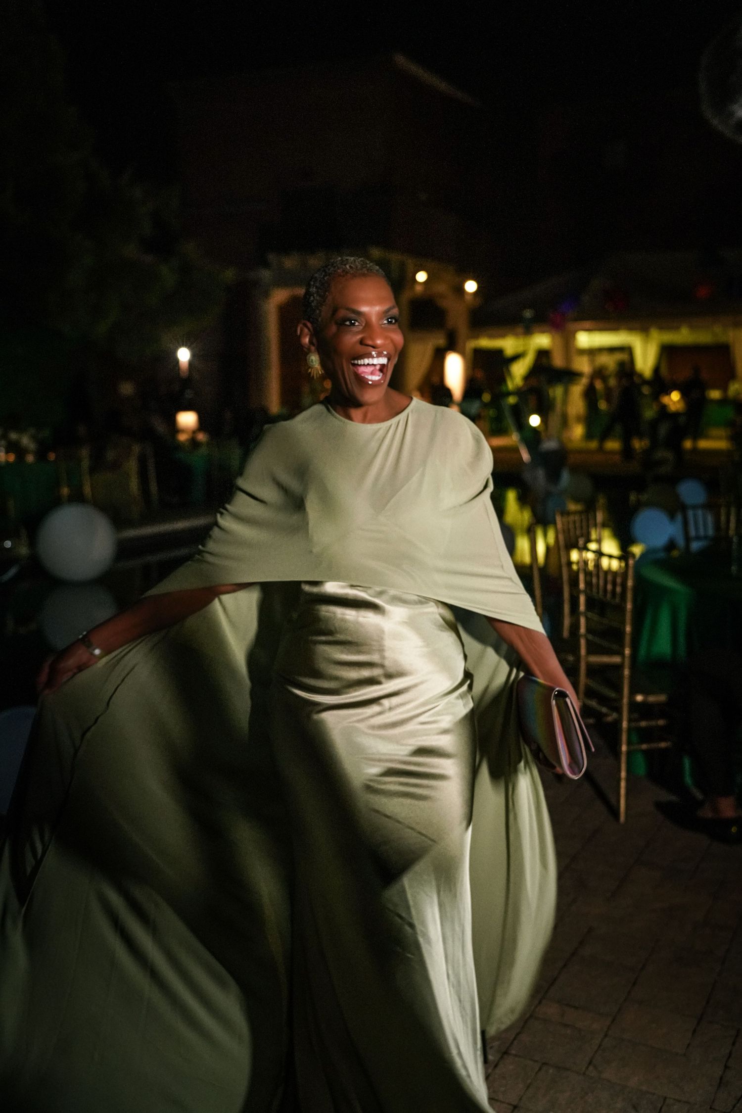 Smiling woman in a flowing sage green satin gown with a cape sleeve twirls joyfully at an outdoor evening event with string lights.