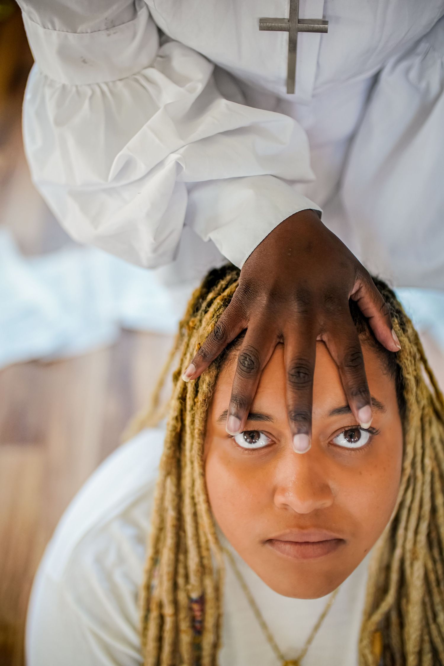 A religious figure in white robes places hands on the head of a person with blonde dreadlocks during a blessing ceremony.
