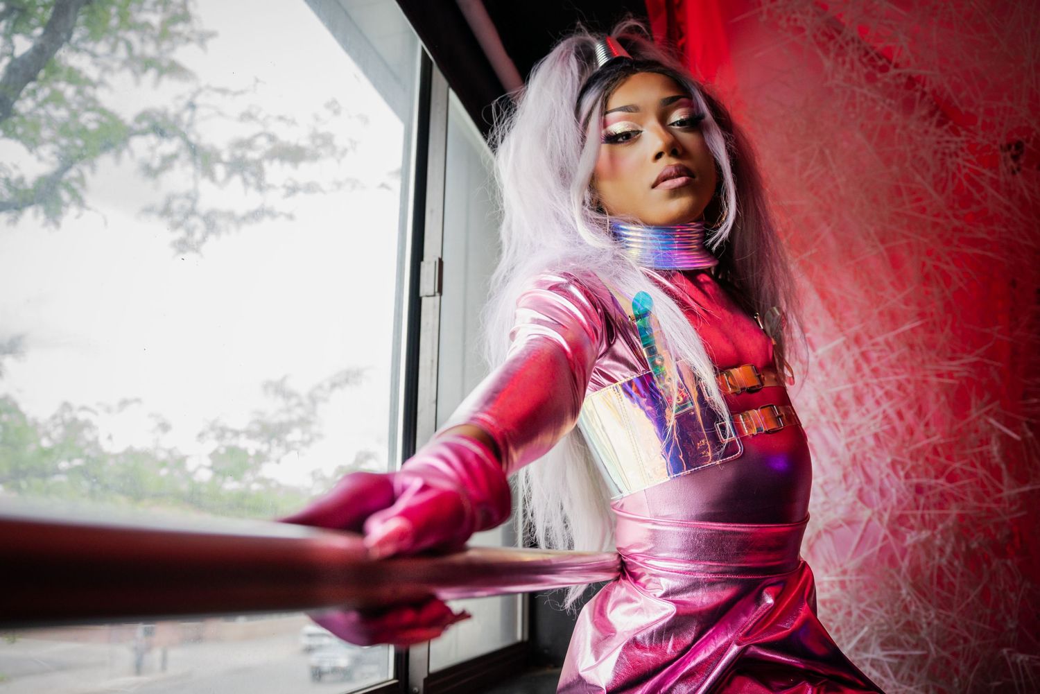 Woman in a shiny pink outfit with pink hair posing near a window with red decorative elements.