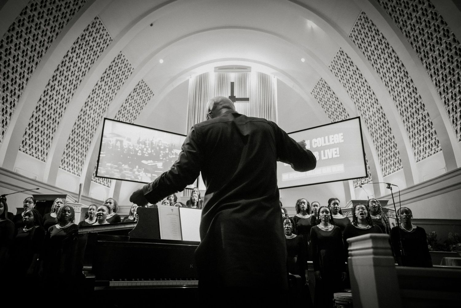 Black and white photo of choir conductor directing large gospel choir beneath decorative church ceiling.