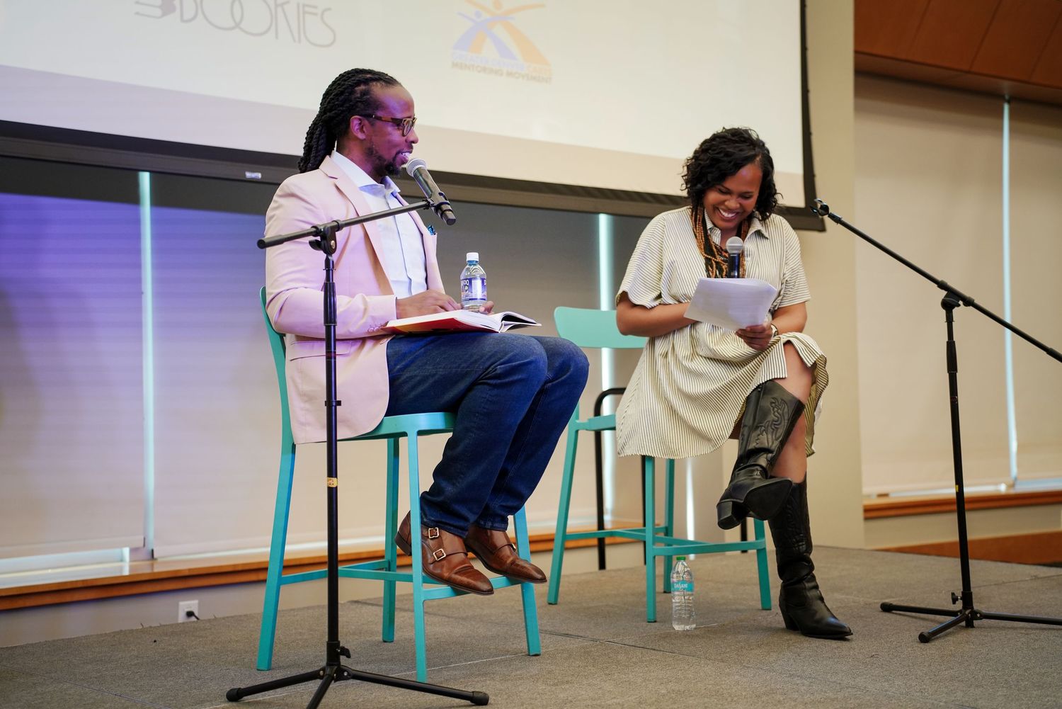 Two speakers sit on teal chairs with microphones during a literary event in front of projection screen.