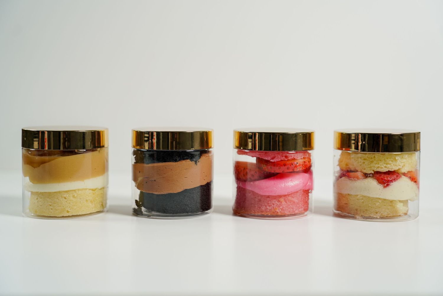 Four clear glass jars with gold lids containing layered colorful desserts on a white background.